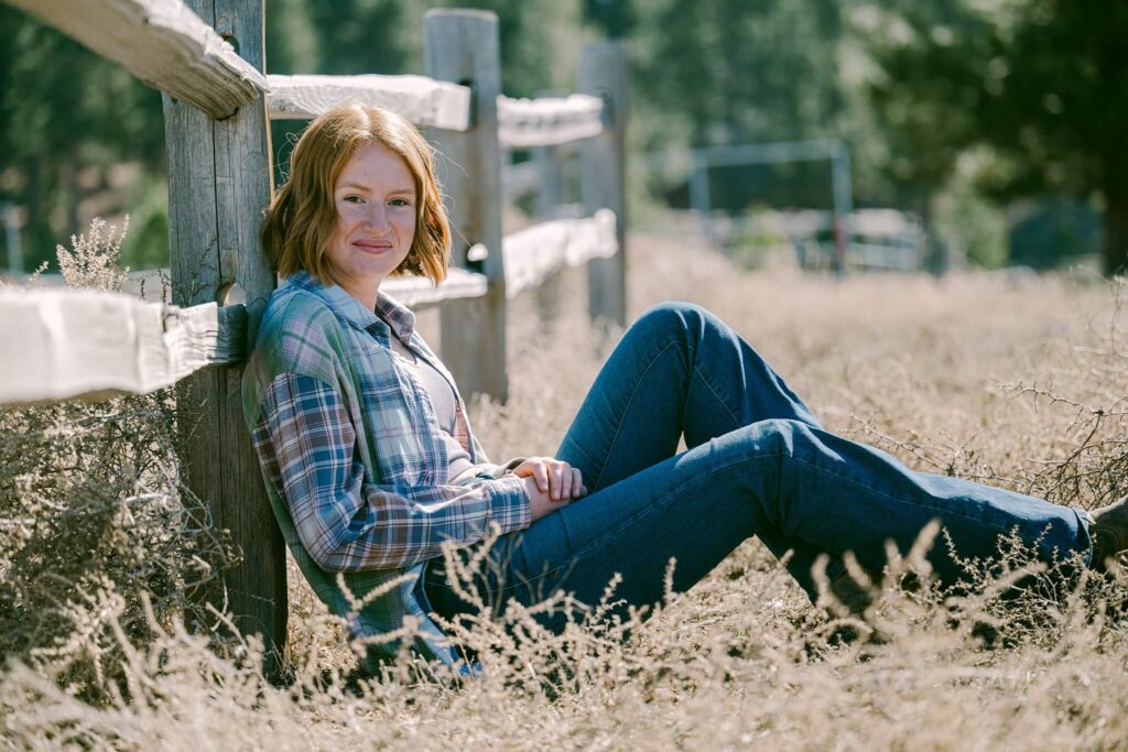 Tahoe senior girl sitting along fence in plaid short looking at camera of Tahoe Senior Photographer Annie X Photographie in Truckee at Piping Rock stables