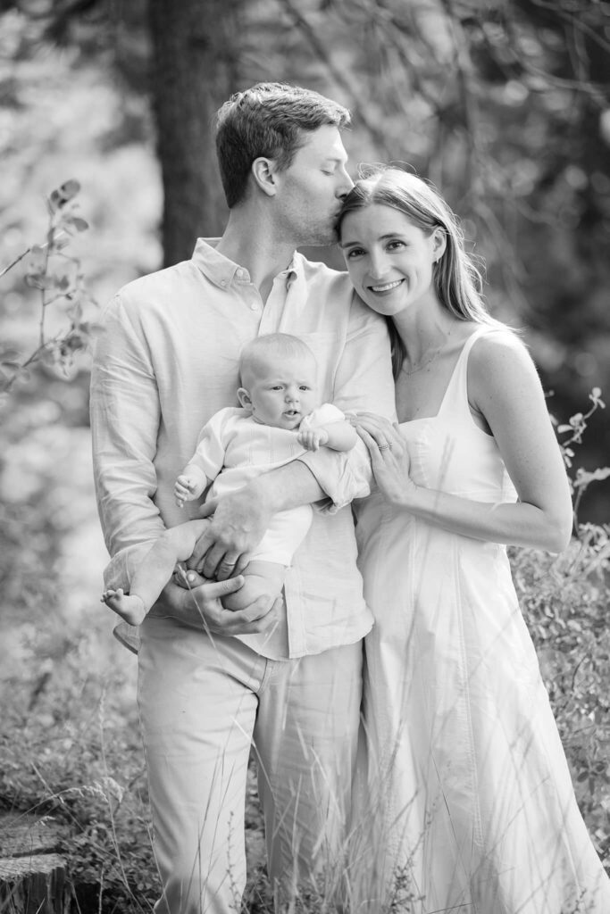 New parents with newborn in black and white dad holding baby and mom looking at camera while dad is kissing her head