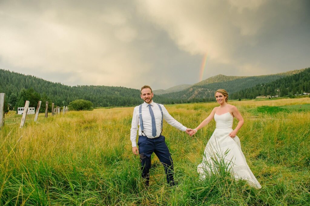 Bride and groom holding hands facing camera with rainbow behind them at Olympic valley stables wedding venue in Palisades Tahoe