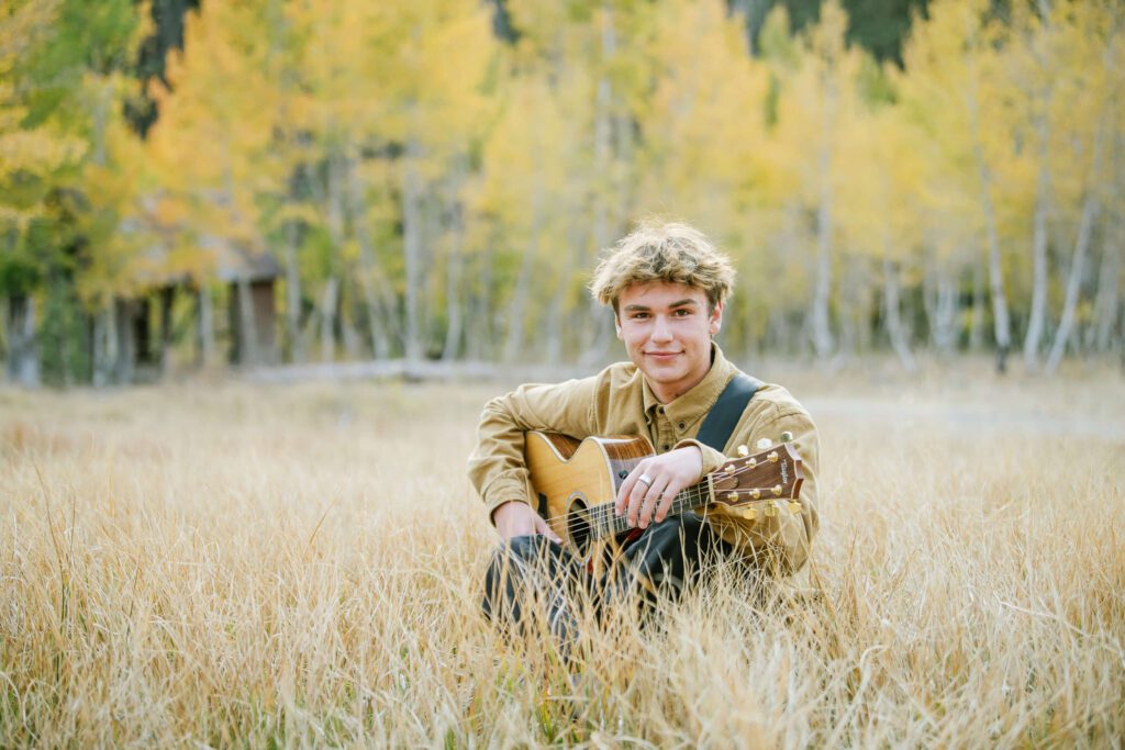 North Tahoe Senior sitting in tall grass and his guitar with fall yellow foliage in teh background captured by Tahoe Senior photographer Annie X Photographie
