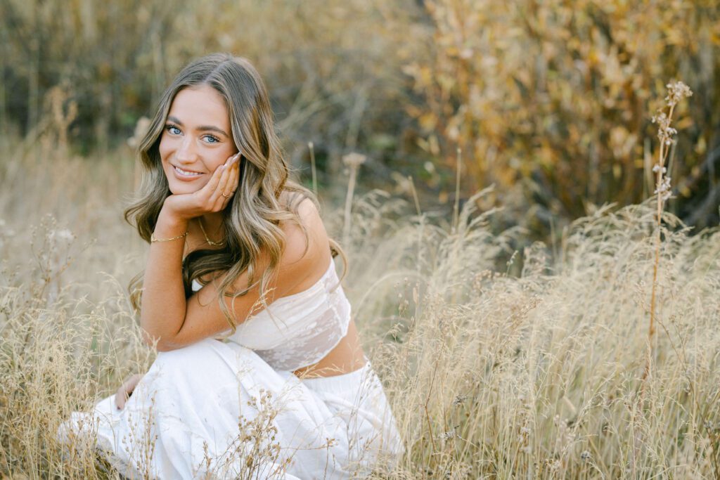 Tahoe Senior portrait by Lake Tahoe senior photographer of a north Tahoe grad sitting in tall grasses in a meadow in the fall wearing a white top and long white skirt.