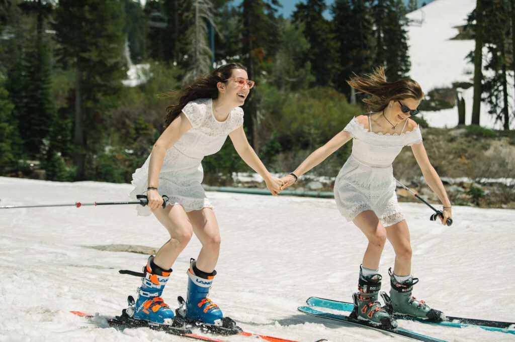 Two Senior girls in white dresses holding hands and skiing down to their graduation from Sugar Bowl academy photographed by Tahoe Senior and family photographer Annie X Photographie