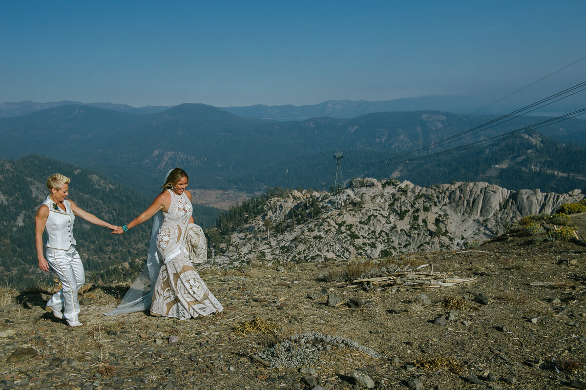 Two brides hand in hand walking at a Lake Tahoe wedding venues High Camp at Palisades Tahoe with views of Lake Tahoe in the distance