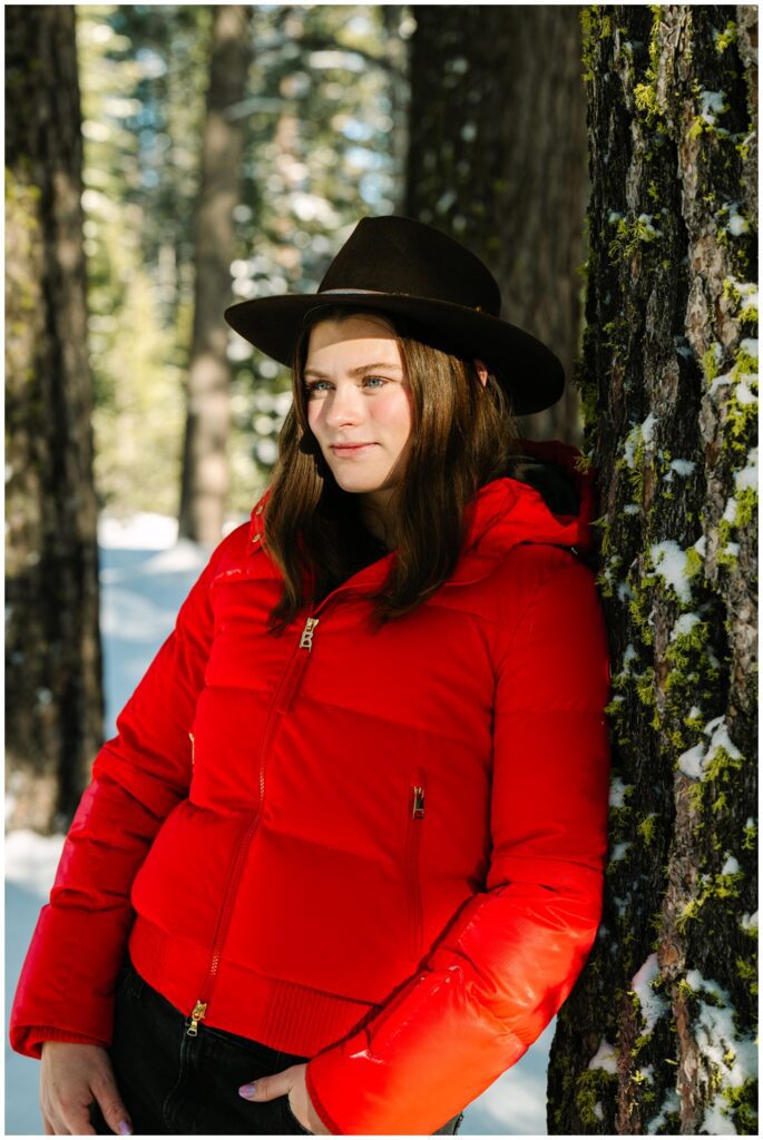 Girl leaning on a mossy tree in a red coat and cowboy hat during a winter portrait session