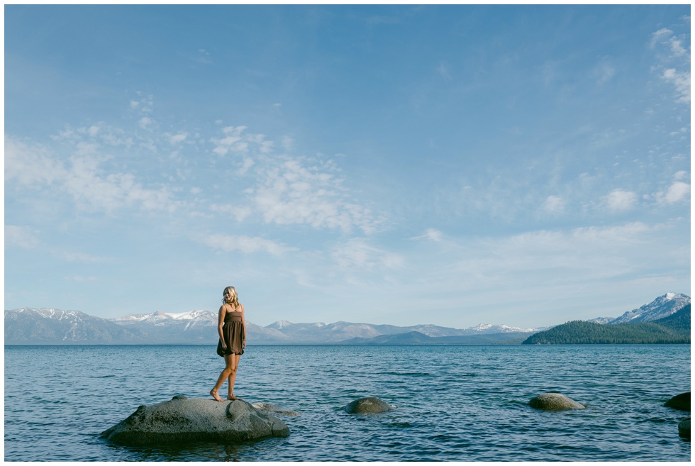 Lake Tahoe senior portraits atmospheric wide portrait of Truckee High School senior in brown mini dress standing alone on granite rock in turquoise Lake Tahoe with expansive snow capped mountain horizon and blue sky Sugar Pine Point spring 2026