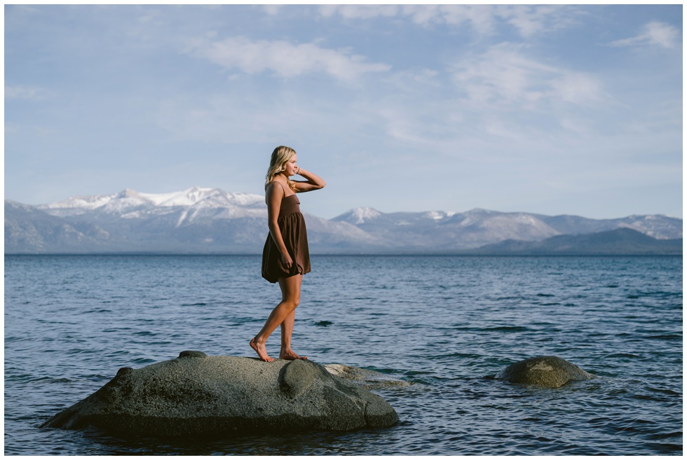 Truckee senior photographer captures Truckee High School senior in brown bubble mini dress standing barefoot on granite lake rock at Sugar Pine Point West Shore with snow capped Sierra Nevada mountains and blue Lake Tahoe behind her spring session