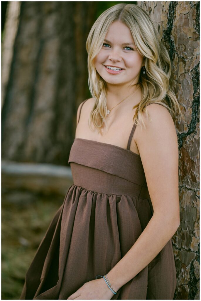 Truckee senior photographer natural light portrait of Class of 2026 senior in brown spaghetti strap dress leaning against Sierra pine tree at Sugar Pine Point State Park joyful expression spring session