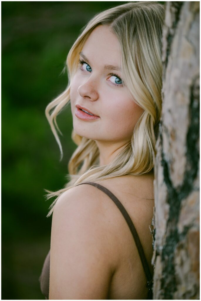 Truckee senior photographer editorial close up of Class of 2026 senior in brown dress with blue eyes and soft expression leaning against ponderosa pine bark Sugar Pine Point natural light spring session