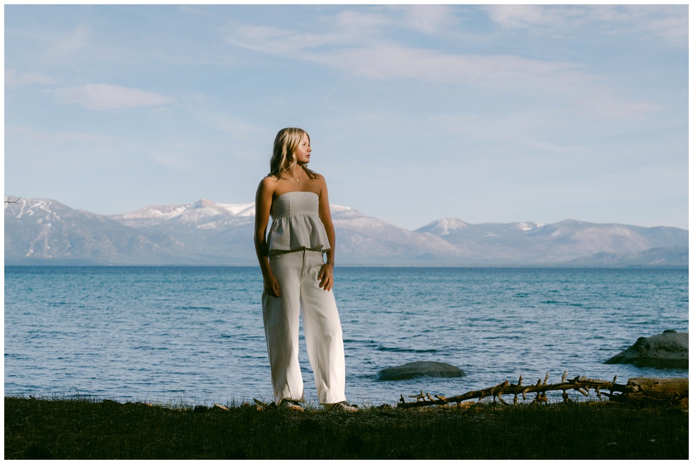 Truckee senior photographer senior girl in grey strapless peplum top and white barrel leg jeans standing at Lake Tahoe West Shore shoreline with snow capped Sierra Nevada mountains and blue lake behind her