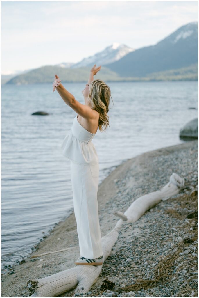 Truckee senior photographer joyful portrait of Class of 2026 senior in white peplum top and wide leg jeans arms raised to sky at Sugar Pine Point lakeside with snow capped Sierra Nevada mountains spring session