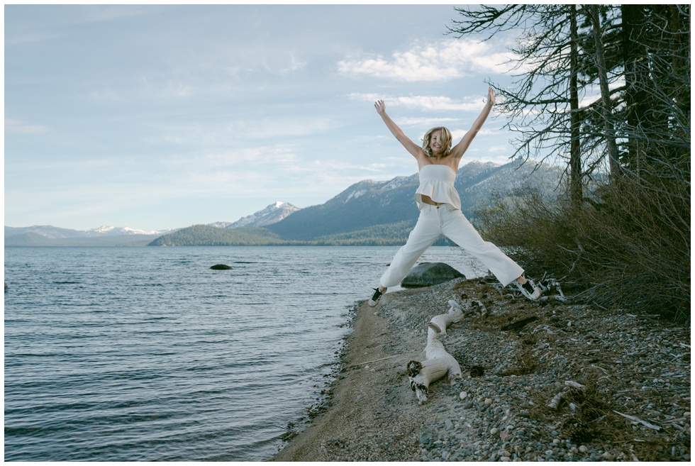 Truckee senior photographer captures joyful jump shot of senior in white outfit on Lake Tahoe West Shore shoreline with snow capped Sierra Nevada mountains and driftwood at Sugar Pine Point