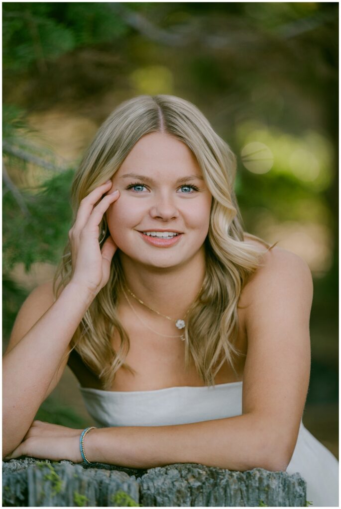 Truckee senior photographer close up portrait of Truckee High School senior in white strapless top smiling with hand to face resting on moss covered pine stump Sugar Pine Point spring 2026