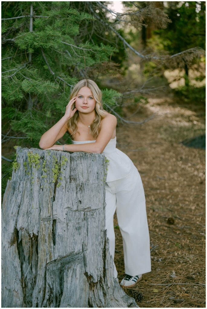 Lake Tahoe senior photos senior girl in white peplum top and wide leg white jeans with Adidas Sambas leaning on weathered mossy pine stump on forest path at Sugar Pine Point