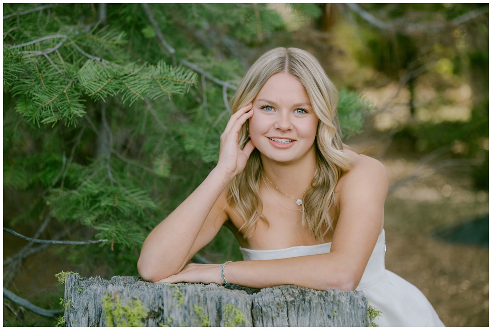 Sugar Pine Point senior portraits spring session senior girl in white strapless top smiling while leaning on moss covered stump surrounded by green pine branches Truckee California