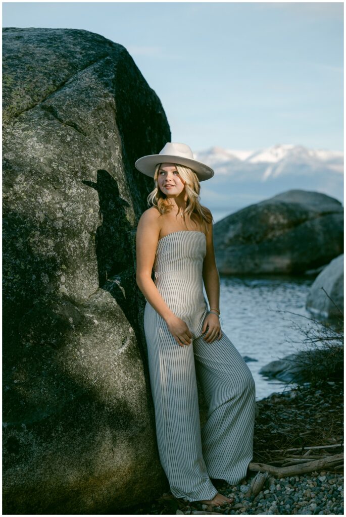 Senior portraits Sugar Pine Point West Shore senior girl in striped strapless jumpsuit and white wide brim hat leaning on granite boulder with soft dusk lake and mountain light behind her