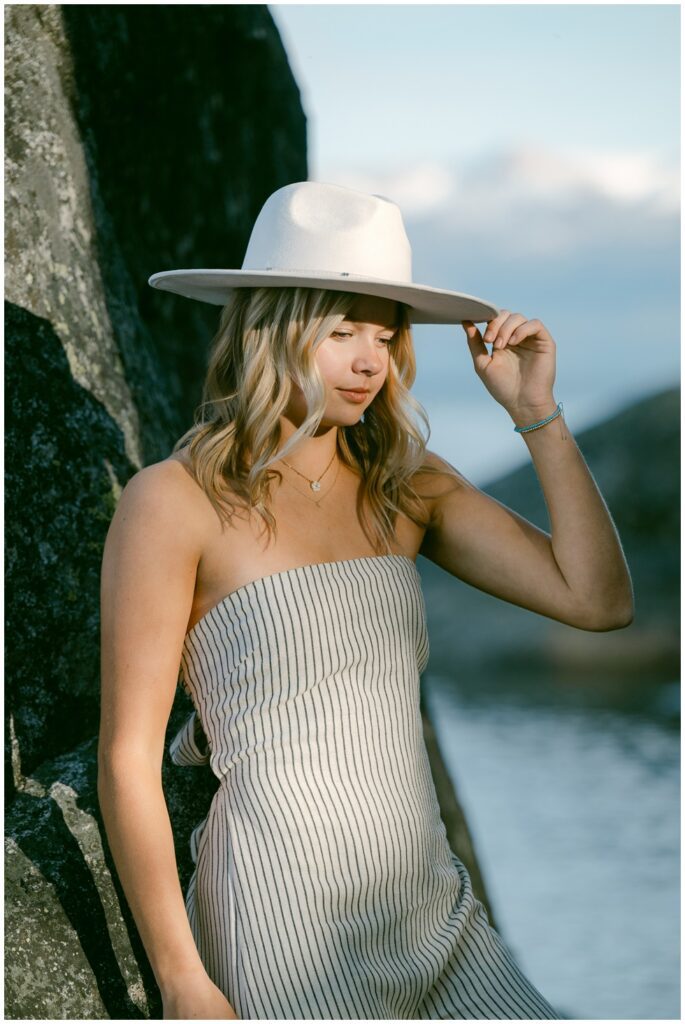 Truckee senior photographer editorial close up of senior girl in striped jumpsuit tipping white hat with warm dramatic lakeside light granite boulder and soft mountain backdrop Sugar Pine Point