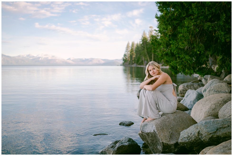 Lake Tahoe senior photos wide environmental portrait of Truckee High School senior in striped strapless jumpsuit sitting on rocks at Sugar Pine Point lakeside with calm lake pine forest and snow capped mountain backdrop spring 2026
