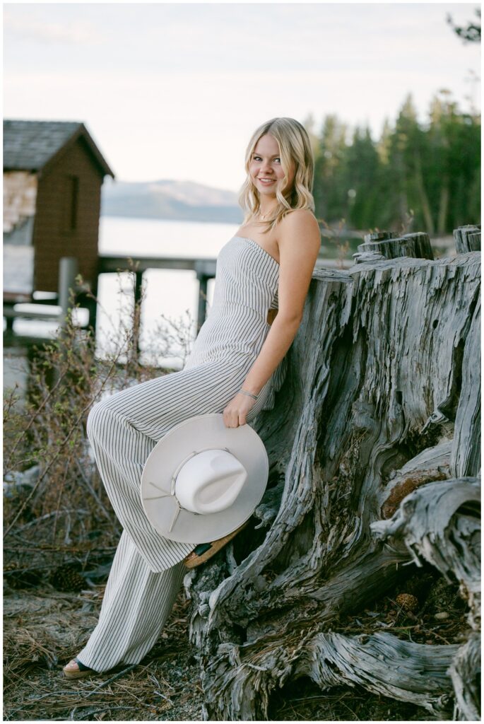 Truckee senior photographer senior girl in striped wide leg jumpsuit sitting on weathered driftwood stump holding white hat at Sugar Pine Point with historic boathouse and Lake Tahoe behind her