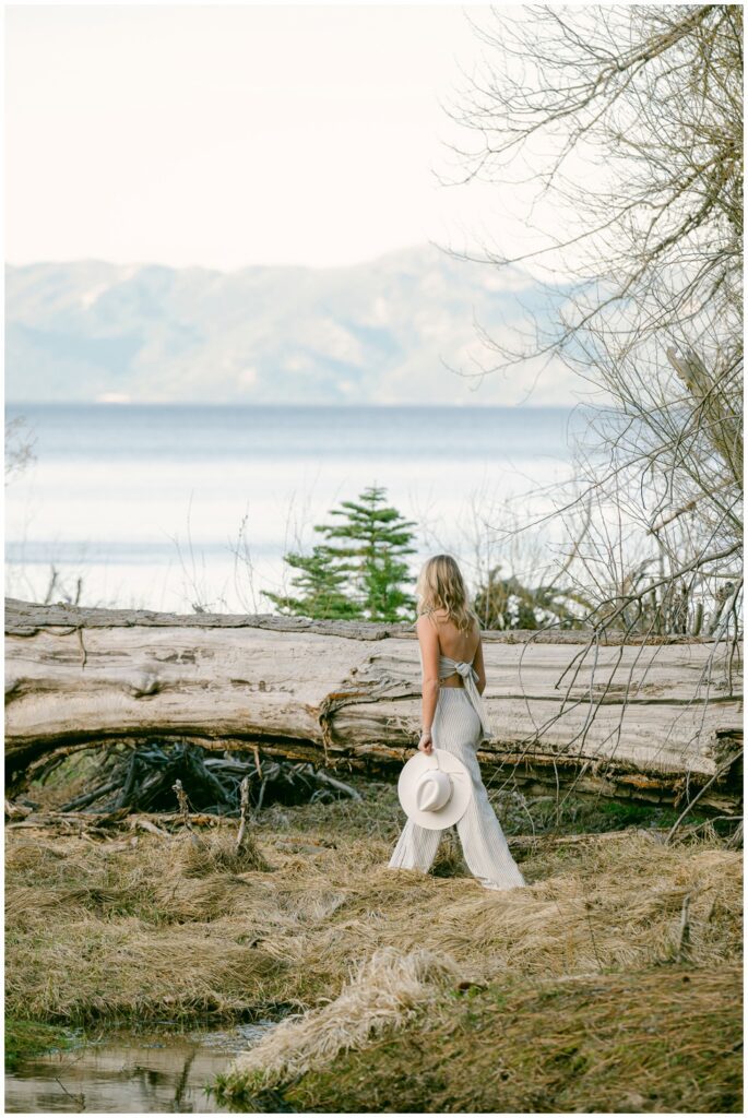West Shore Lake Tahoe senior portraits atmospheric back portrait of senior girl in open back striped jumpsuit holding white hat standing by driftwood log at Sugar Pine Point with calm lake and mountain backdrop