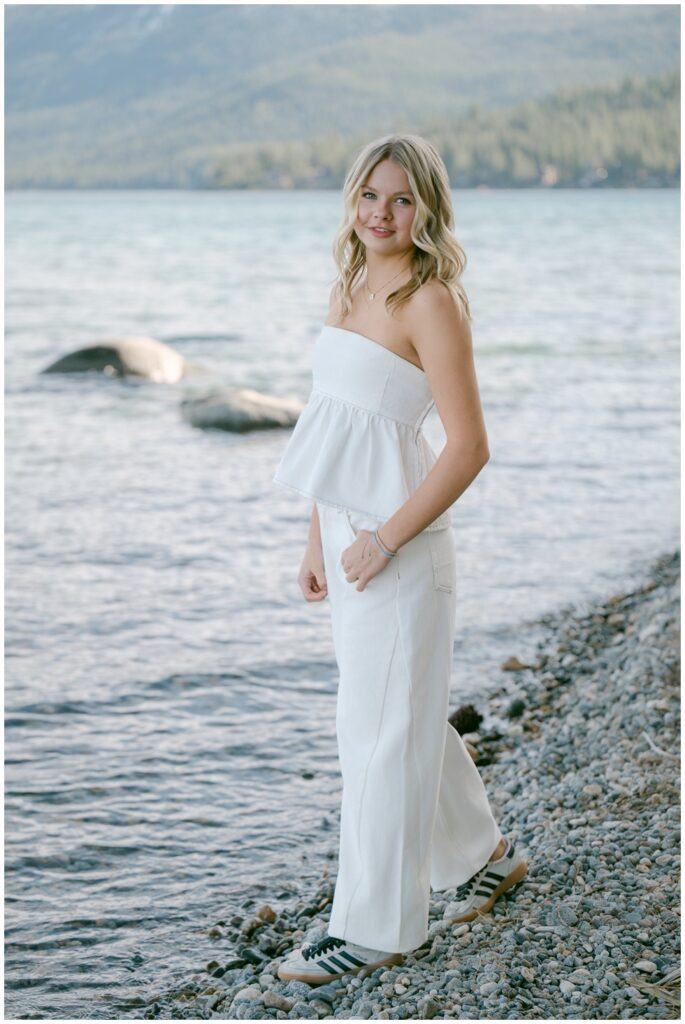 West Shore Lake Tahoe senior photos full body portrait of Class of 2026 senior in white tonal peplum top and barrel leg jeans on rocky Sugar Pine Point shoreline with turquoise lake and green mountain backdrop