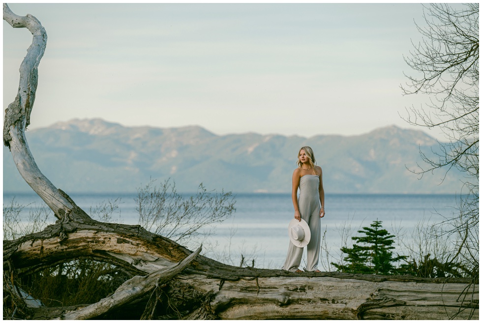 Truckee senior photographer editorial environmental portrait of Truckee High School Class of 2026 senior in striped jumpsuit holding white hat standing on weathered driftwood log at Lake Tahoe West Shore with full Sierra Nevada mountain panorama and soft spring light